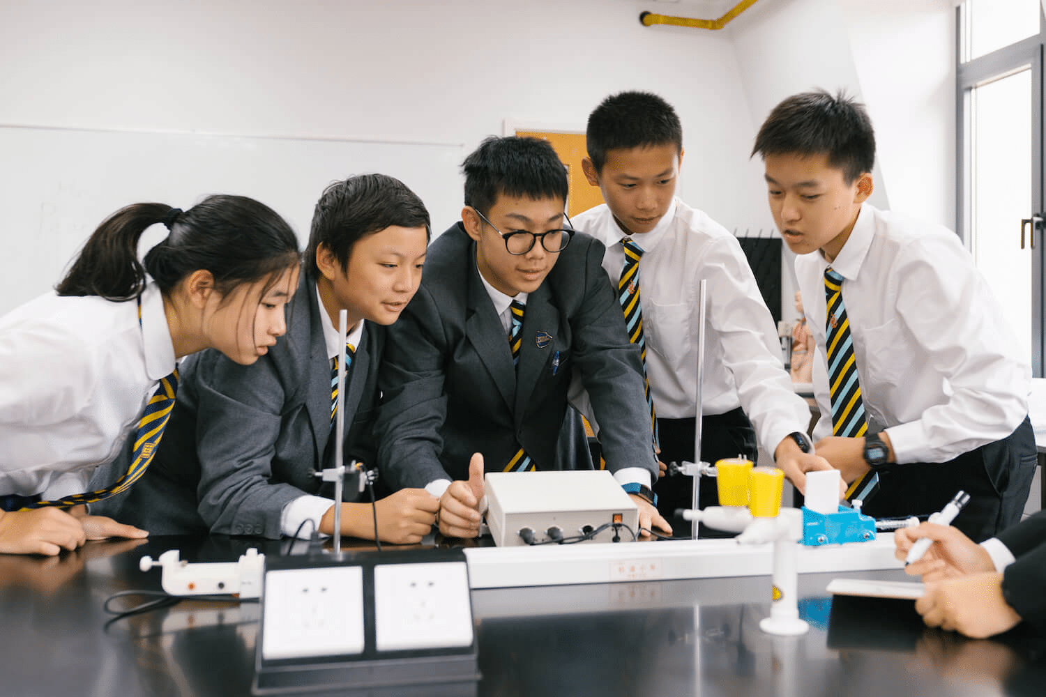 Five students in school uniforms from an international education group gather around a science lab bench, engaged in a practical experiment with lab equipment, working together and observing the set-up attentively.