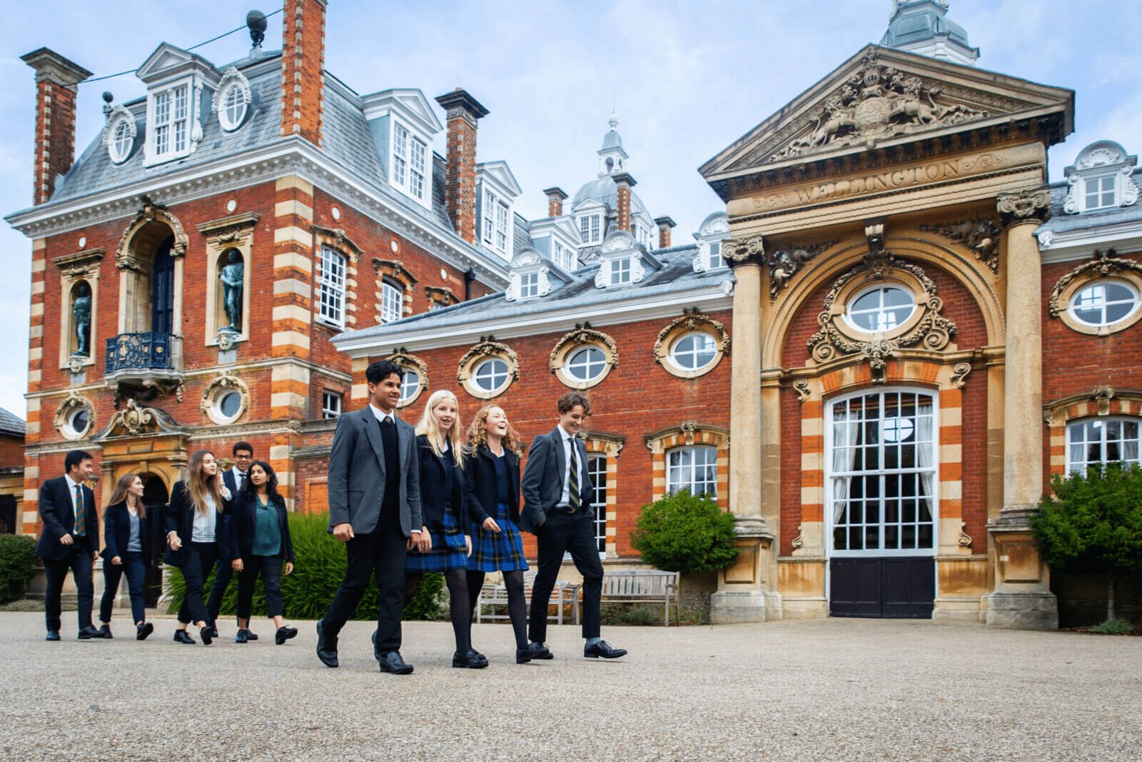A group of students in school uniforms walk together outside a large, ornate red-brick building, part of an international school network. The arched windows and classical details shine under a clear sky, reflecting a bright learning environment.