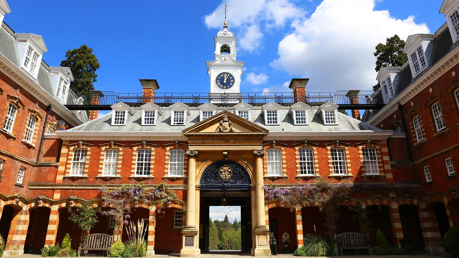 A grand, historic brick building with a clock tower, arched windows, and a columned entrance stands amid greenery beneath a partly cloudy blue sky—an inspiring centrepiece for the international schools alliance.
