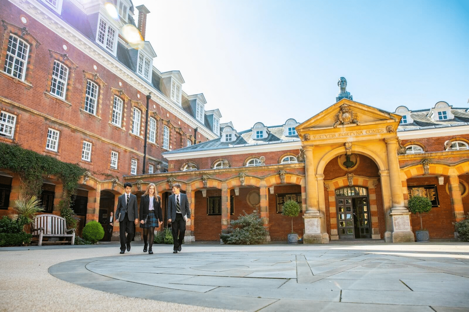 Four people in formal attire walk together in the courtyard of a historic brick building, home to an international school group, with arched entrances and large windows on a bright, sunny day.